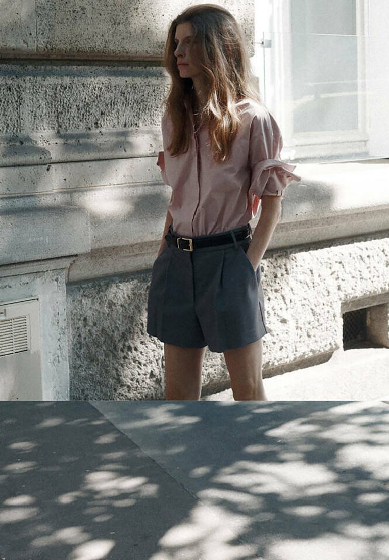 Femme aux longs cheveux portant un t-shirt rose et un short foncé, debout les mains dans les poches près d'un mur en pierre, sous une lumière douce du jour et des motifs d'ombre.