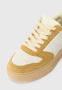 Sneaker med overdel i creme læder og tan ruskind, tykke creme snørebånd, perforeret tåboks og en tekstureret beige gummisål.