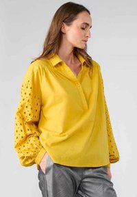 Blusa gialla con scollatura a bottoni e maniche texture caratterizzate da motivi intagliati. Realizzata in tessuto leggero.