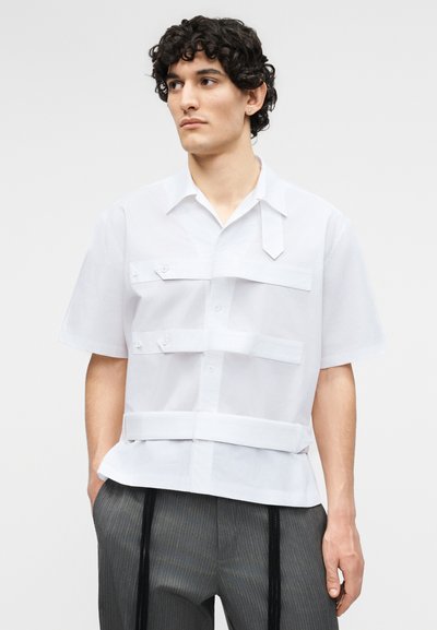 Νέος άνδρας με σγουρά μαλλιά που φοράει λευκό πουκάμισο με κοντά μανίκια και τιράντες και γκρι παντελόνι με λεπτές ρίγες, στέκεται με τα χέρια στις τσέπες.