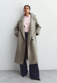 Lång grå kappa i ull, med en bred krage, dubbelknäppt design och sidofickor, bärs över en ljusrosa stickad topp och mörka jeans.