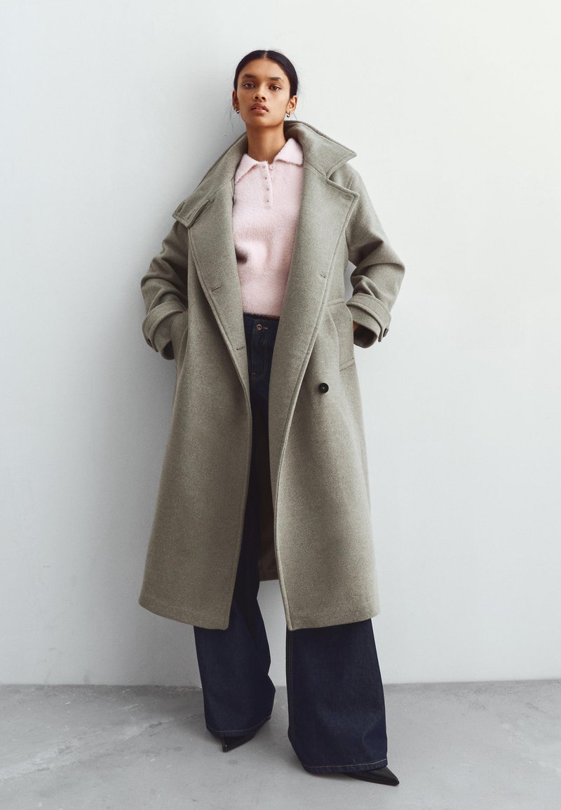 Lång grå kappa i ull, med en bred krage, dubbelknäppt design och sidofickor, bärs över en ljusrosa stickad topp och mörka jeans.