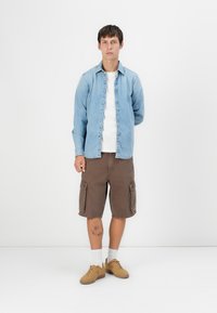 Lichtblauwe spijkerblouse over een wit T-shirt, gecombineerd met bruine cargoshorts. Tan sneakers en witte sokken maken de outfit compleet.
