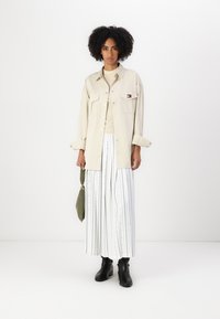 Beige skjorte med knapper og to lommer, kombinert med hvite vide bukser med striper og farget prikker. Svart ankelstøvler og grønn veske.
