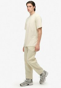 Beige katoenen t-shirt met korte mouwen, gecombineerd met bijpassende beige broek. Draagt grijze sportschoenen met zwarte accenten.