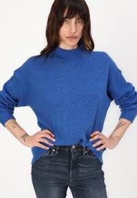 Modra pletena majica z visokim ovratnikom, ohlapnega kroja in rebrasto teksturo, v kombinaciji s temnimi kavbojkami, ki prikazujejo gumbe in žepe.