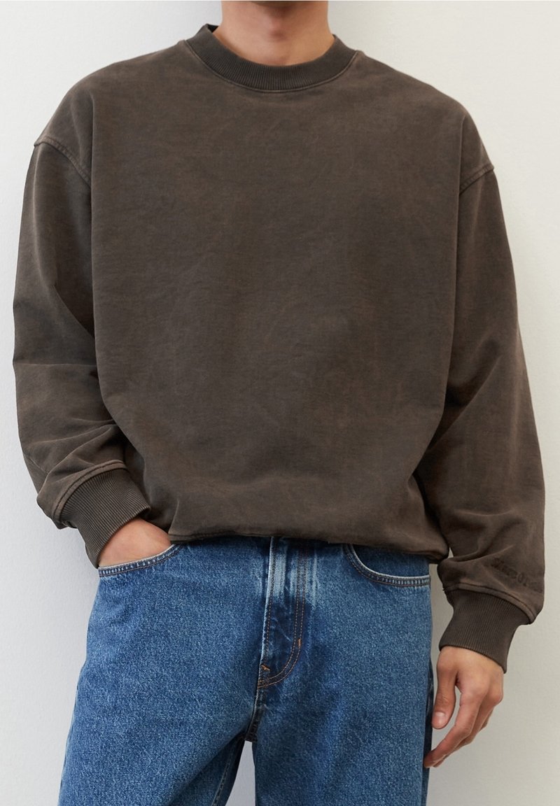 Personne portant un sweat-shirt marron foncé ample avec poignets et col côtelés, associé à un jean denim bleu classique, debout avec une main dans la poche.
