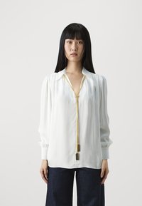 Blusa bianca con scollatura profonda e dettaglio della cerniera dorata. Presenta maniche lunghe con polsini arricciati. Abbinata a pantaloni in denim scuro.