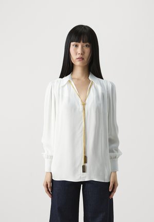 Blusa bianca con scollatura profonda e dettaglio della cerniera dorata. Presenta maniche lunghe con polsini arricciati. Abbinata a pantaloni in denim scuro.
