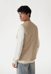 Beige gesteppte Weste mit glatter Textur, Rundhalsausschnitt und ärmellosem Design. Über einem weißen Langarmshirt und hellblauen Jeans getragen.
