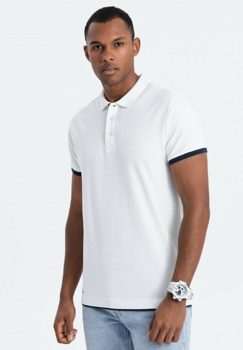 Polo bianco con colletto a due bottoni, rifiniture blu navy sui maniche e tessuto strutturato. Il modello indossa jeans azzurri e un orologio bianco.