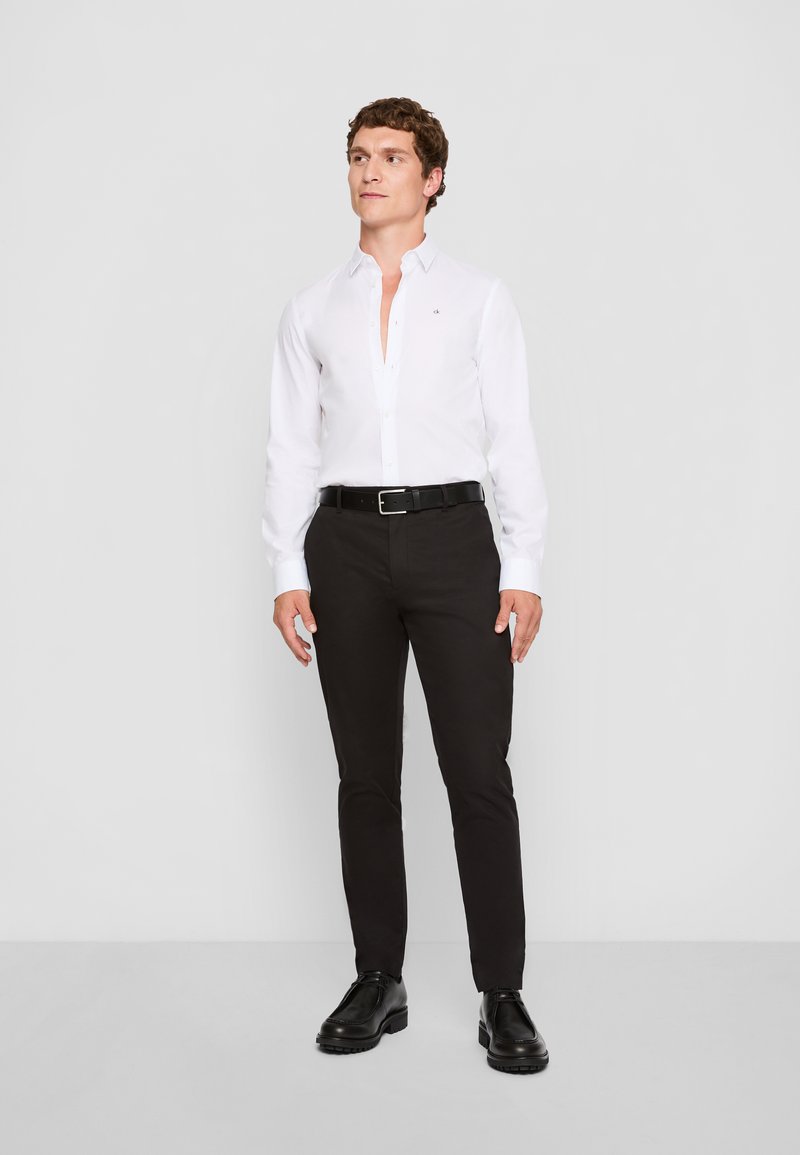 Weißes Langarmhemd mit Kragen, schwarze Hose und schwarze Schuhe. Das Hemd hat ein kleines Logo auf der Brust. Glattes Gewebe, taillierte Passform.