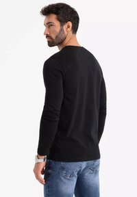 Černá košile s dlouhými rukávy, kulatým výstřihem a slim fit, v kombinaci s světle modrými džíny. Na sobě ji má muž s krátkými tmavými vlasy a vousy.
