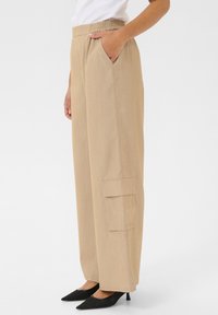 Pantalons larges beiges avec une taille élastique, poches latérales et détails de poche cargo. Fabriqués en tissu texturé, associés à des escarpins noirs à bout pointu.