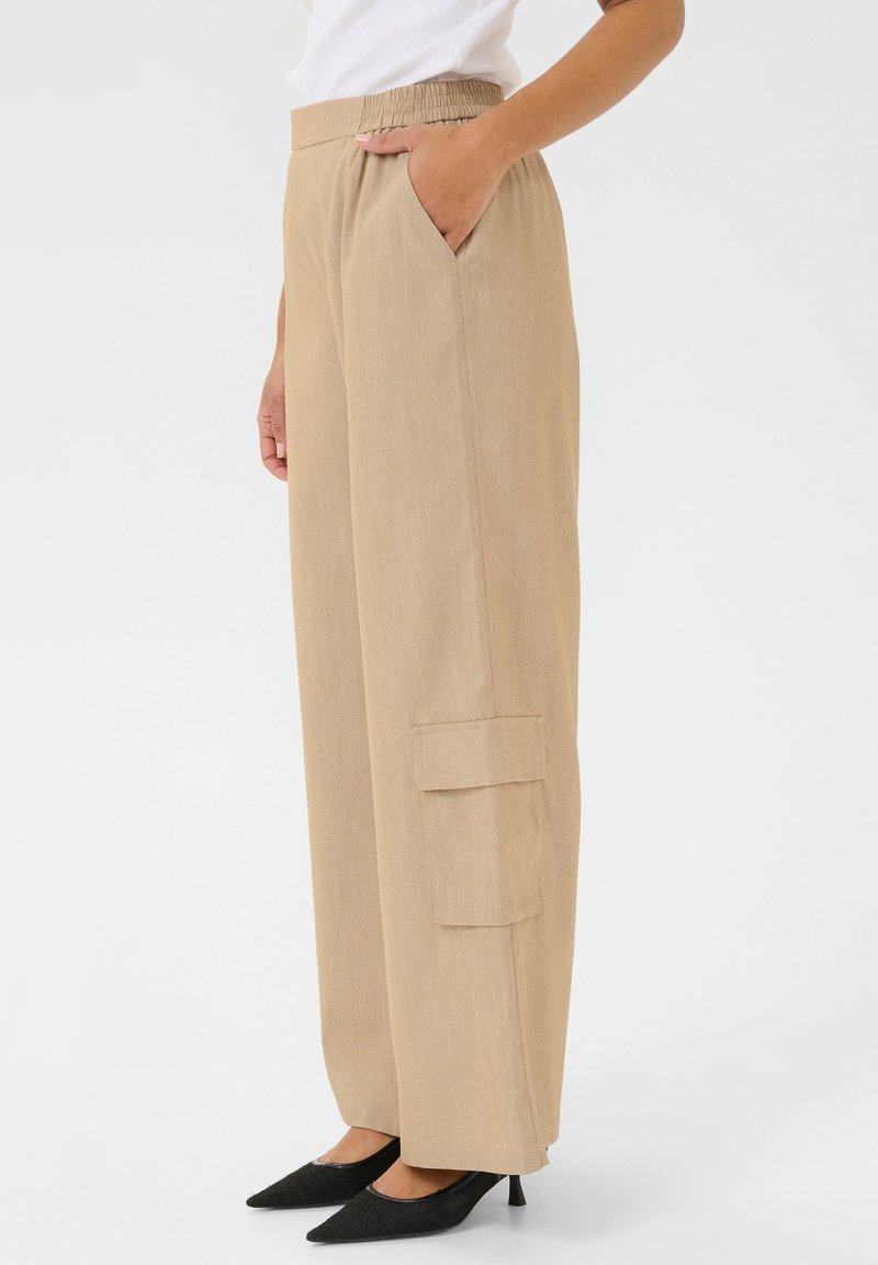 Pantalons larges beiges avec une taille élastique, poches latérales et détails de poche cargo. Fabriqués en tissu texturé, associés à des escarpins noirs à bout pointu.