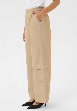 Personne portant un pantalon beige à jambes larges avec poche cargo et ceinture élastique, associé à des talons noirs pointus et un haut blanc.