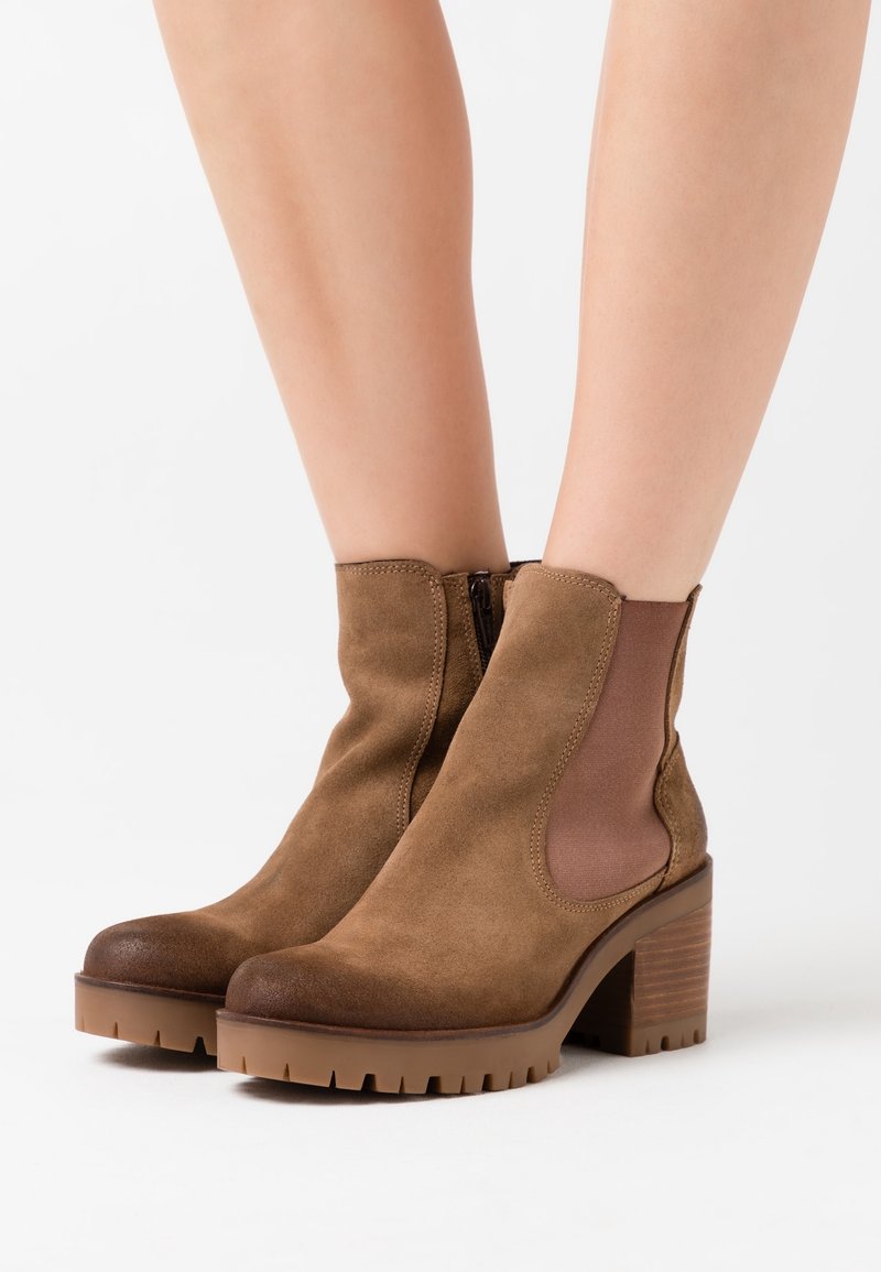 Bottes en daim marron montant à la cheville avec talons épais, panneaux élastiques latéraux et semelles en caoutchouc texturé, portées sur des jambes nues sur fond blanc.