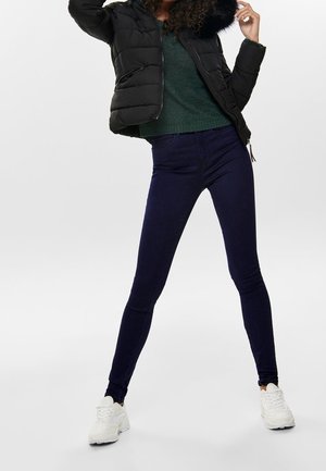 Woman wearing black puffer jacket with fur hood, green sweater, dark skinny pants, and white sneakers standing with legs apart on white background.