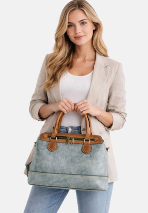 Vrouw met lang blond haar, die een beige blazer en een wit topje draagt, en een grote blauwe handtas vasthoudt met bruine leren handvatten.