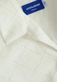 Tessuto di cotone bianco con un motivo a rombi e dettagli ritagliati. È visibile un'etichetta blu con la scritta "JACK & JONES ORIGINALS".