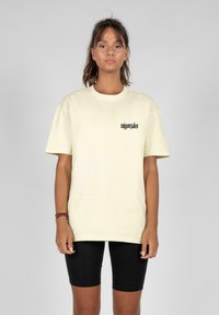 Camiseta de algodón amarilla clara con mangas cortas, cuello redondo y logo de texto negro en el lado izquierdo. Se lleva con pantalones cortos de ciclismo negros.