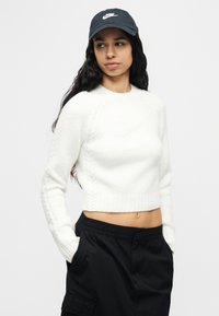 Γυναίκα με μακριά μαύρα μαλλιά, που φοράει μαύρο καπέλο Nike, λευκό κοντό πουλόβερ και μαύρο παντελόνι, με τα χέρια στις τσέπες, κοιτάζοντας αριστερά.