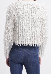 Vit texturerad tröja med en fransmönster, kort design och långa ärmar. Ili klädd med mörkblå tighta jeans.