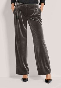 Vrouw in donkergrijze wijde fluwelen broek met plooien, zwarte puntige hakken, en handen in de zakken tegen een lichte achtergrond.