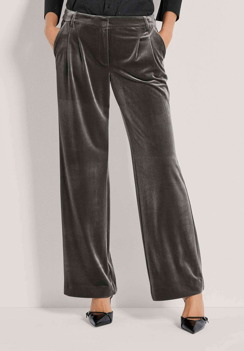 Vrouw in donkergrijze wijde fluwelen broek met plooien, zwarte puntige hakken, en handen in de zakken tegen een lichte achtergrond.