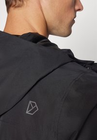 Juoda gobtuvu striukė su lygiu paviršiumi. Netoli peties yra geometrinis logotipas šviesesniu atspalviu. Paprastas dizainas, be išskirtinių raštų.