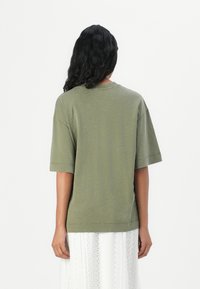 Olijfgroen t-shirt met korte mouwen, losse pasvorm met een geribbelde ronde hals en een zachte textuur, gezien van de achterkant, gecombineerd met een witte gemusterde rok.