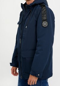 Tmavě modrá zateplená bunda s kapucí, předním zipem, dvěma velkými kapsami a texturovaným ramenním popruhem s kruhovým logem.
