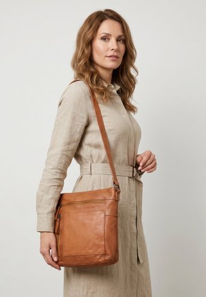 Vrouw met golvend lichtbruin haar die een beige jurk met ceintuur draagt en een grote lichtbruine leren schoudertas met voorvakken met rits meeneemt.
