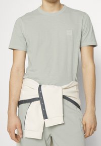 Ljusgrön kortärmad t-shirt med en texturerad finish, ihop med en krämfärgad tröja knuten runt midjan. Diskret logotypdetalj på t-shirten.