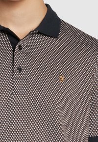 Gros plan sur un polo avec un col noir, des boutons noirs et un petit emblème brodé jaune sur un tissu à motifs.