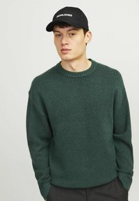 Dark green knitted sweater with ribbed crew neckline and long sleeves, worn over black pants, paired with a black cap featuring white logo.