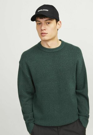 Dark green knitted sweater with ribbed crew neckline and long sleeves, worn over black pants, paired with a black cap featuring white logo.