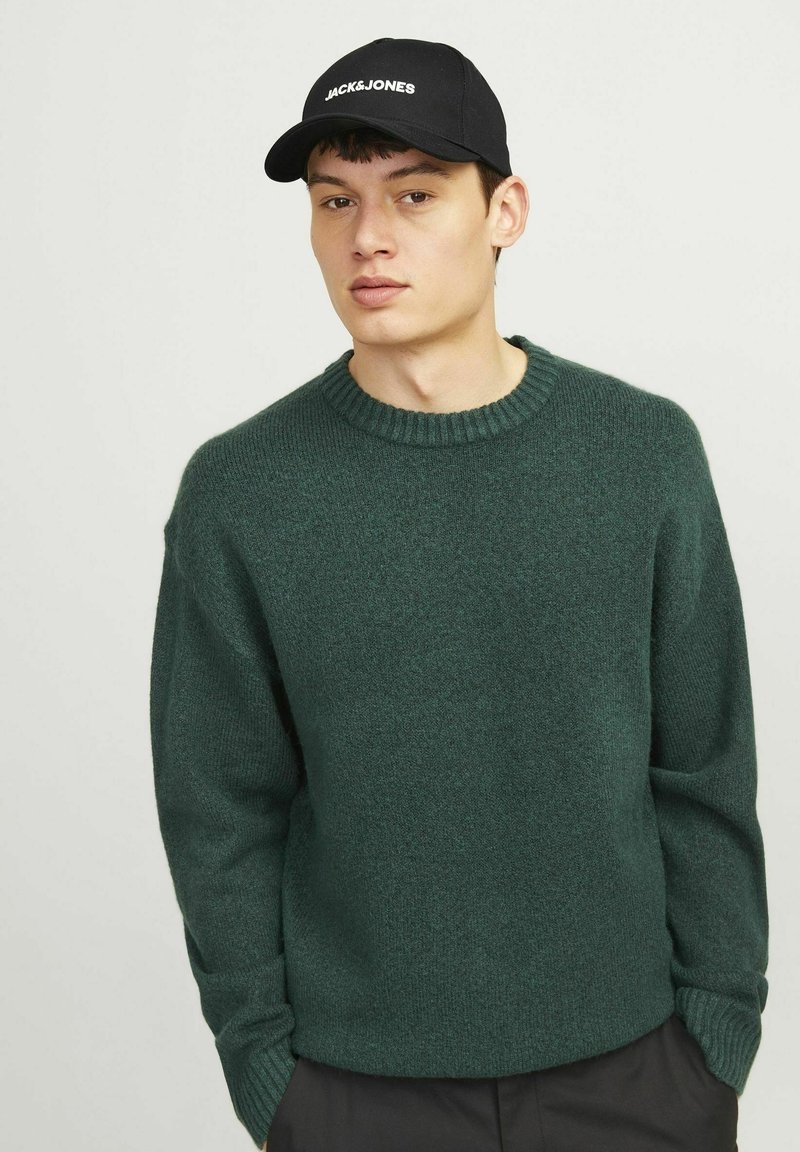 Dark green knitted sweater with ribbed crew neckline and long sleeves, worn over black pants, paired with a black cap featuring white logo.