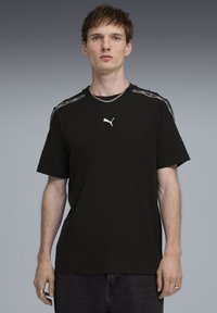 Νεαρός άνδρας που φοράει μαύρο T-shirt Puma με λογότυπο στο στήθος και λευκή επένδυση στους ώμους, στέκεται μπροστά σε φόντο με γκρι διαβάθμιση.