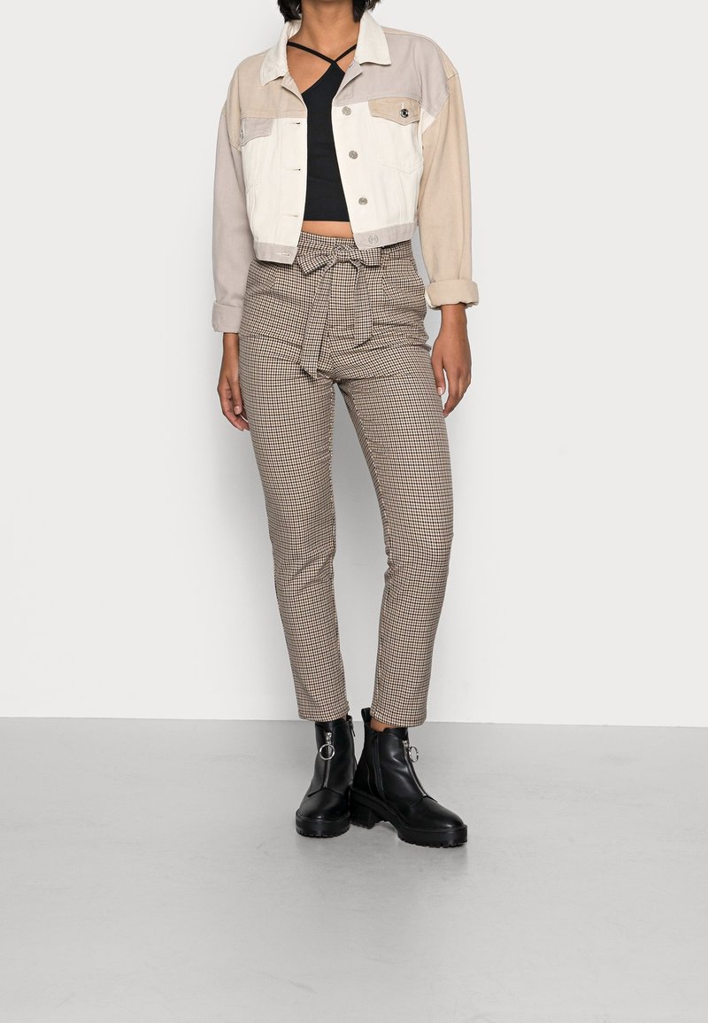 Personne portant une veste courte beige et blanche, un crop top noir, un pantalon marron à carreaux taille haute avec ceinture à nouer et des bottines noires.