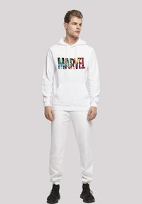 Balts kapucis ar krāsainu "MARVEL" grafiku, kurā attēloti komiksu varoņi. Apvienots ar atbilstošām baltām sporta biksēm un melnām kedām.