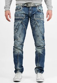 Jean en denim bleu avec un lavage décoloré, motifs de couture uniques et écussons de marque sur les poches avant, associés à des baskets blanches.