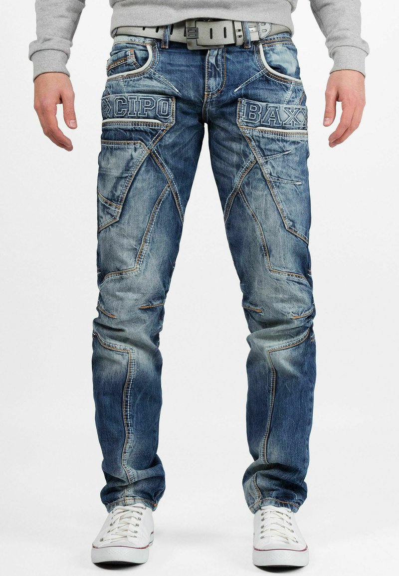 Jean en denim bleu avec un lavage décoloré, motifs de couture uniques et écussons de marque sur les poches avant, associés à des baskets blanches.