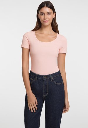 Light pink short-sleeve bodysuit with a scoop neckline featuring "GUESS JEANS" printed in gold. Worn with dark denim high-waisted jeans.