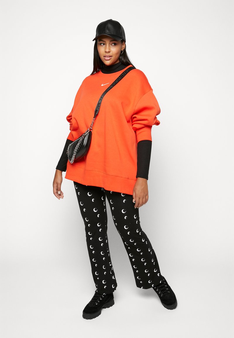 Sweatshirt orange surdimensionné, col roulé noir à côtes en dessous, pantalon noir avec motif de croissant de lune blanc, et chaussures noires.