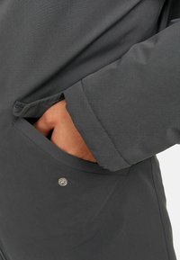 Indumento esterno grigio scuro con una texture liscia e opaca, dotato di una tasca laterale e di un dettaglio a scatto metallico vicino all'orlo. Polsino regolabile.