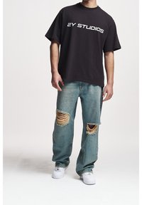 Černé oversized tričko s nápisem "2Y STUDIOS" v bílé barvě. Opotřebované modré džíny s ošoupanými trhlinami na kolenou. Bílé tenisky doplňují outfit.