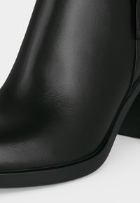 Gros plan d'une botte noire en cuir lisse à la cheville, avec un bout arrondi et un talon carré sur fond blanc.