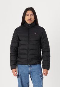 Zwarte pufferjack met hoge kraag, ritsluiting en gestikte panelen. Draagt over blauwe denim, met een klein logo-embleem op de borst.