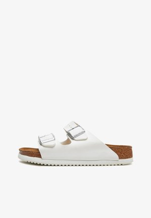 White sandals featuring two adjustable buckles, cork footbed, textured rubber outsole, and a minimalist design with a smooth finish.